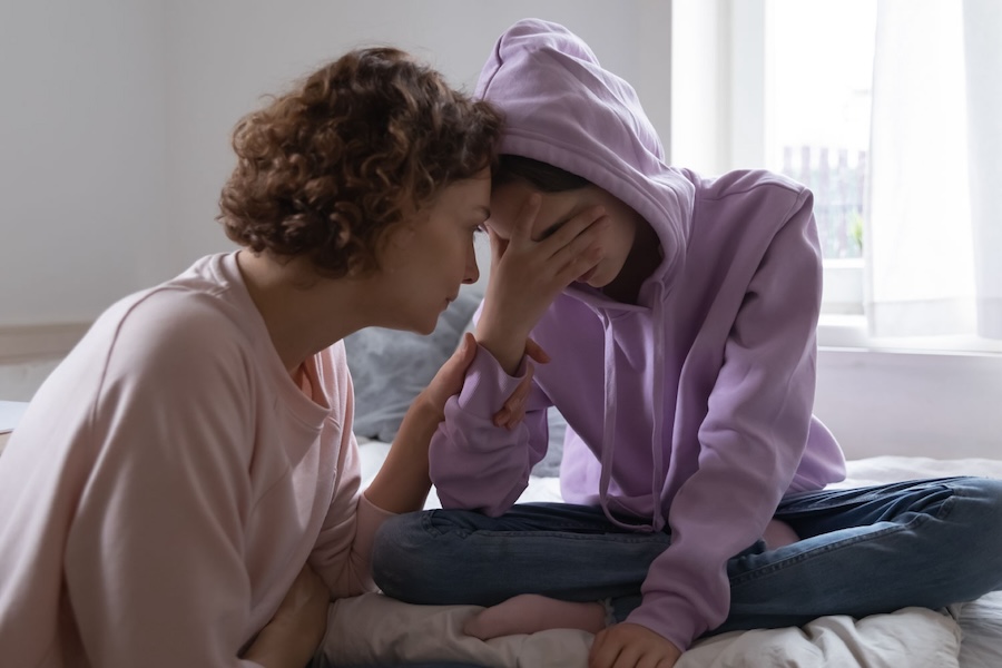 Image of a mother and daughter sitting on a bed while the mom holds the daughters face, she's upset