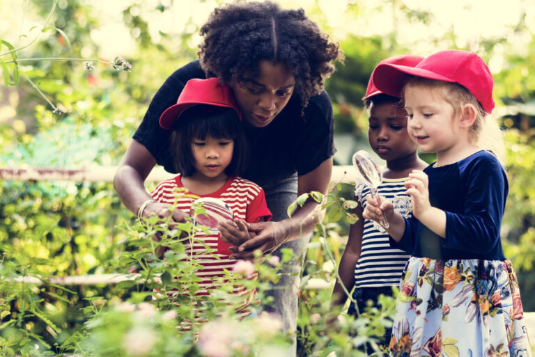 A woman and three children outside in a gardening looking at plants with magnified glasses.