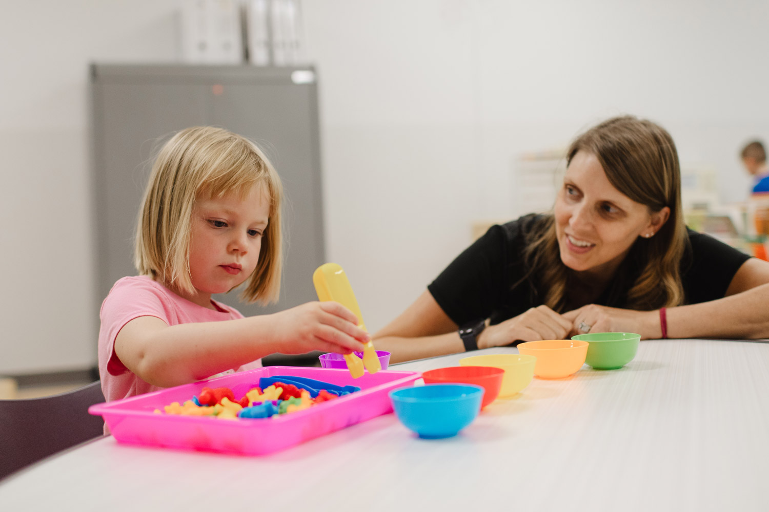 Photo of a FamilyForward staff member looking at a young girl as we places colorful objects into the colored bowls.