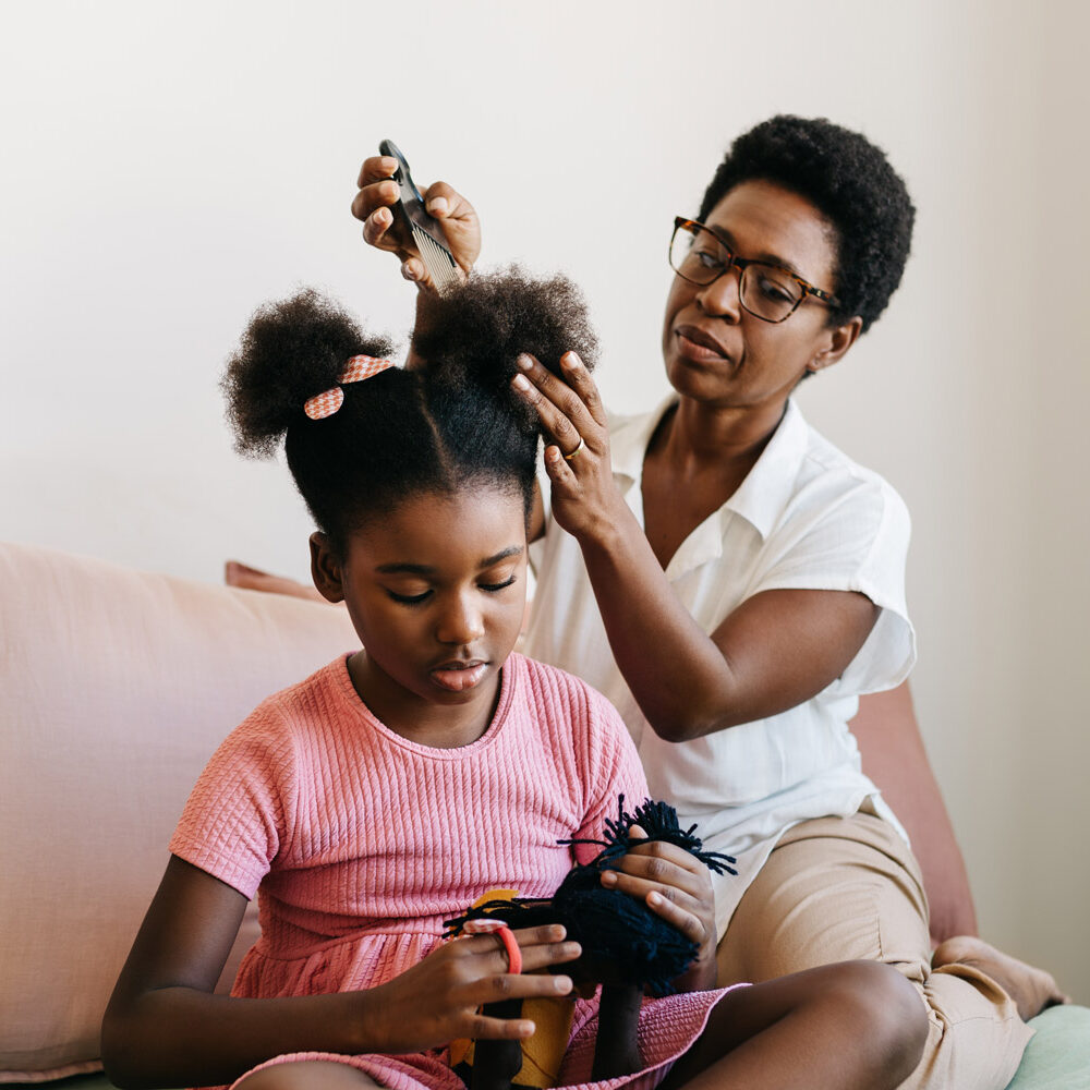 An older black woman combing out a young black girl's hair softly with a hair pick.