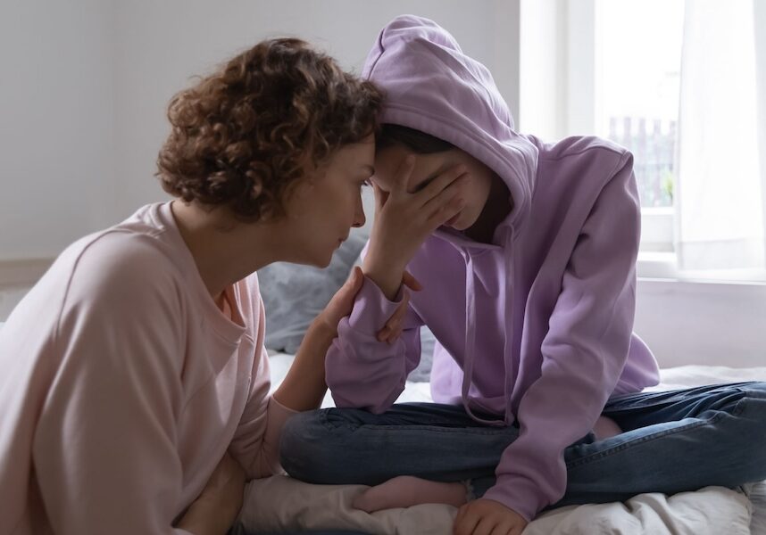 Image of a mother and daughter sitting on a bed while the mom holds the daughters face, she's upset