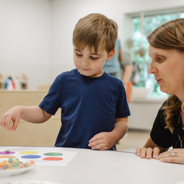 A woman kneeling beside a young boy as he places fruit loops onto a paper with the corresponding colors.