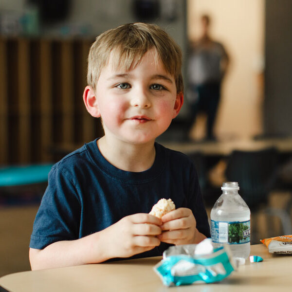 A young boy softly smiling and eating a snack at a desk.