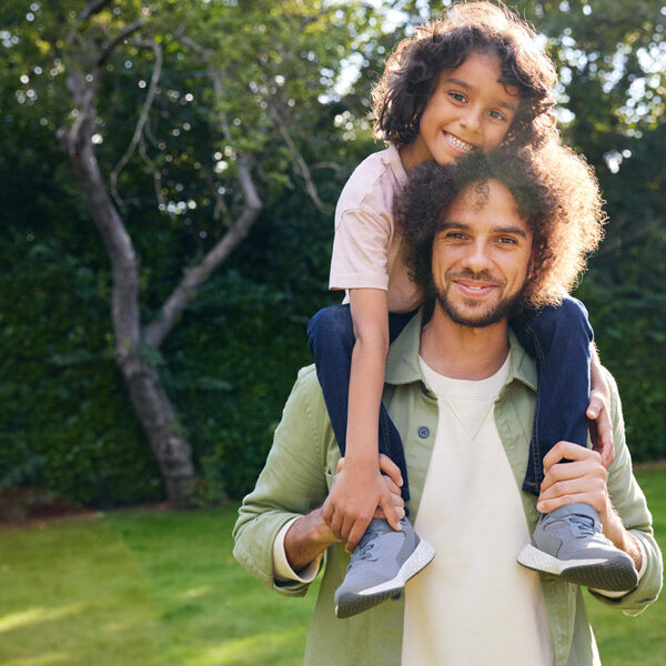 Father Carrying Son On Shoulders As They Walk In Garden