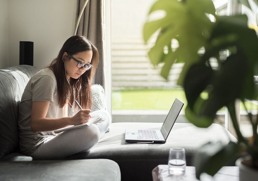 A young woman sitting on a couch doing work with headphones in.